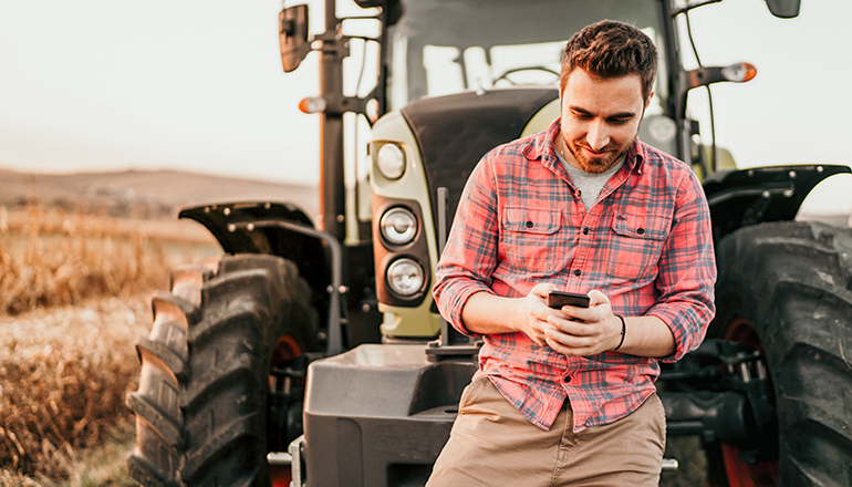 Young man holding phone standing by Tractor in field