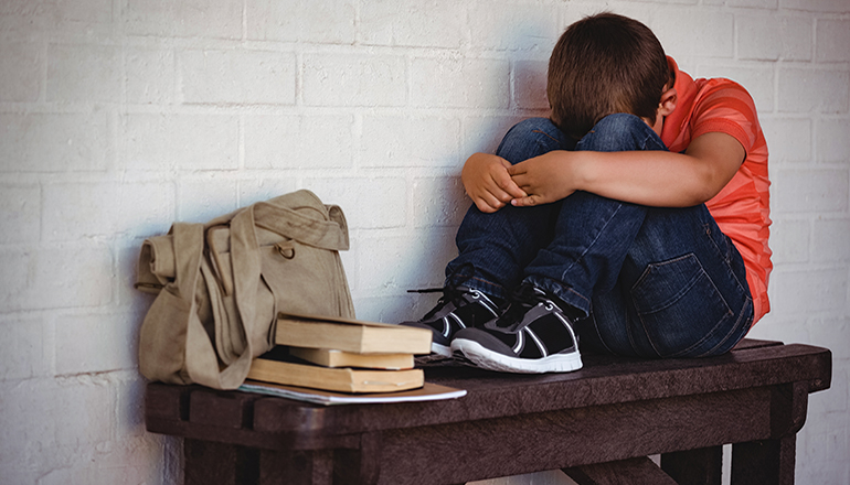 Unhappy boy or child sitting on bench by wall with books (school)